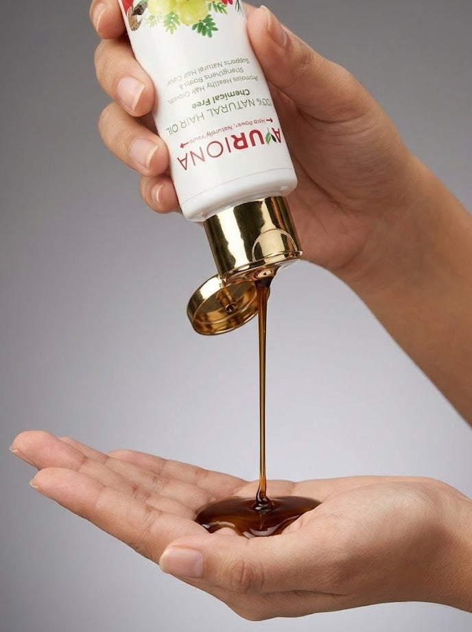 Hand holding a bottle of Ayuriona product with oil being poured onto another hand against a gray background